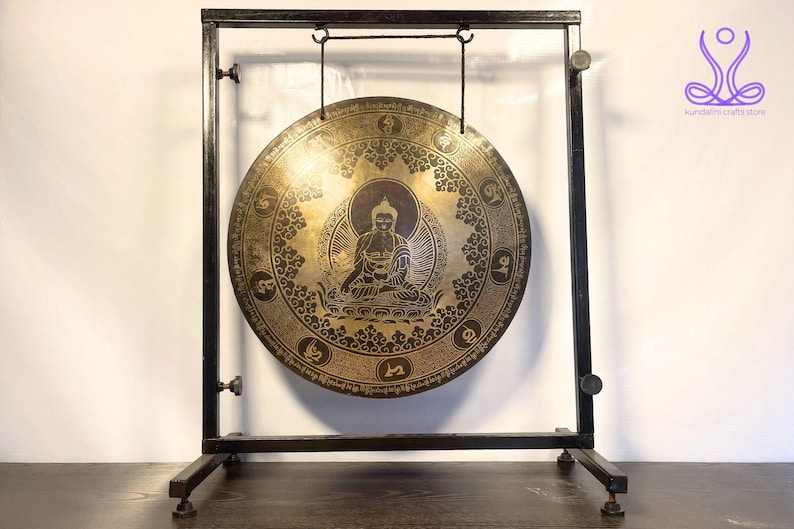 K&ouml;nnte beinhalten: Ein gro&szlig;er, runder, goldfarbener Gong mit aufwendigen Designs und einer Buddha-Figur in der Mitte, der an einem schwarzen Metallst&auml;nder h&auml;ngt.