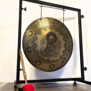 K&ouml;nnte beinhalten: Ein gro&szlig;er, runder, Messinggong mit einem schwarzen Metallst&auml;nder und einem Holzhammer. Der Gong hat ein detailliertes Design mit einer sitzenden Buddha-Figur, die von komplizierten Mustern umgeben ist.