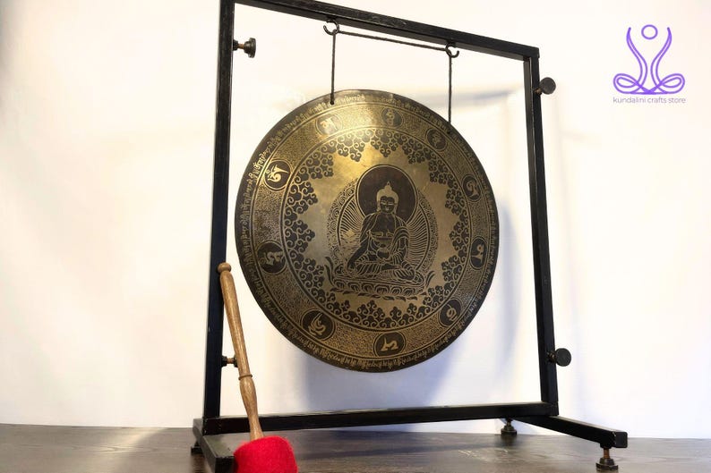 K&ouml;nnte beinhalten: Ein gro&szlig;er, runder, Messinggong mit einem schwarzen Metallst&auml;nder und einem Holzhammer. Der Gong hat ein detailliertes Design mit einer sitzenden Buddha-Figur, die von komplizierten Mustern umgeben ist.