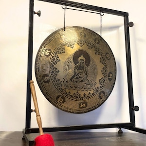 K&ouml;nnte beinhalten: Ein gro&szlig;er, runder, goldfarbener Gong mit einem schwarzen Metallst&auml;nder und einem Holzhammer. Der Gong zeigt ein detailliertes Design einer sitzenden Buddha-Figur, umgeben von komplizierten Mustern und Symbolen.