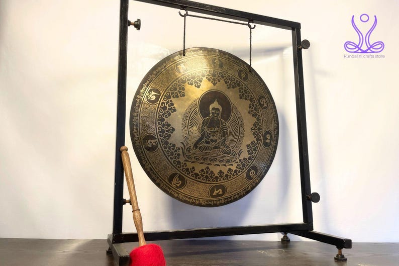 K&ouml;nnte beinhalten: Ein gro&szlig;er, runder, goldfarbener Gong mit einem schwarzen Metallst&auml;nder und einem Holzhammer. Der Gong zeigt ein detailliertes Design einer sitzenden Buddha-Figur, umgeben von komplizierten Mustern und Symbolen.