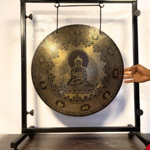 K&ouml;nnte beinhalten: Ein gro&szlig;er, runder, goldfarbener Gong mit aufwendigen Verzierungen und einer Darstellung von Buddha in der Mitte. Der Gong h&auml;ngt an einem St&auml;nder mit einem schwarzen Metallrahmen.