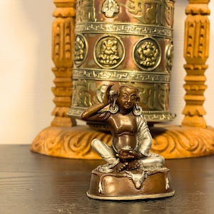 May include: A bronze-toned statue of a seated figure with one hand raised to their ear, set before a decorative prayer wheel. The statue is on a brown base. The prayer wheel is made of copper and wood.
