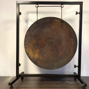 K&ouml;nnte beinhalten: Ein gro&szlig;er, runder, antik aussehender Gong, der an einem schwarzen Metallst&auml;nder h&auml;ngt. Der Gong ist aus Metall und hat eine dunkelbraune Patina. Der St&auml;nder ist aus schwarzem Metall und hat ein schlichtes Design.