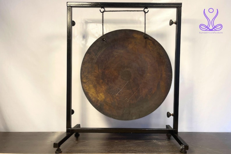 K&ouml;nnte beinhalten: Ein gro&szlig;er, runder, antik aussehender Gong, der an einem schwarzen Metallst&auml;nder h&auml;ngt. Der Gong ist aus Metall und hat eine dunkelbraune Patina. Der St&auml;nder ist aus schwarzem Metall und hat ein schlichtes Design.