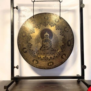 K&ouml;nnte beinhalten: Ein gro&szlig;er, runder, goldfarbener Gong mit aufwendigen Verzierungen und einer Darstellung von Buddha in der Mitte. Der Gong h&auml;ngt an einem schwarzen Metallst&auml;nder.