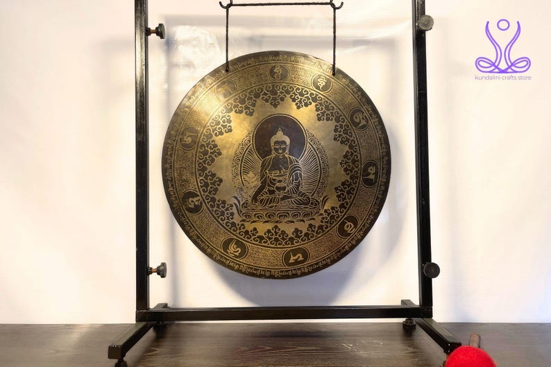 K&ouml;nnte beinhalten: Ein gro&szlig;er, runder, goldfarbener Gong mit aufwendigen Verzierungen und einer Darstellung von Buddha in der Mitte. Der Gong h&auml;ngt an einem schwarzen Metallst&auml;nder.
