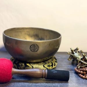 May include: A traditional Tibetan singing bowl with a brass finish and a decorative design on the bottom. The bowl is resting on a cushion with gold embroidery. A wooden striker with a red felt tip and a set of wooden beads are also included.