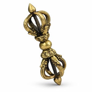 Puede incluir: Un objeto ritual vajra de metal dorado con una empuñadura esférica central flanqueada por dos extremos simétricos. Cada extremo consta de cuatro puntas curvas que se unen en un ápice, con detalles en relieve en la base de las puntas.