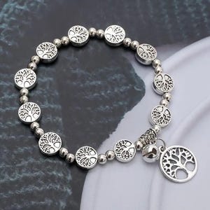 May include: Silver-toned bracelet featuring a tree of life design. The bracelet has round, flat charms with tree engravings, connected by small silver beads. A dangling charm with a tree design and a small bell adds detail.