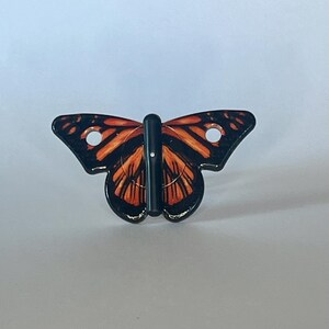 May include: A black and orange butterfly-shaped hook with two holes on the wings. The hook is made of plastic and has a black metal rod.