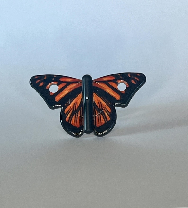 May include: A black and orange butterfly-shaped hook with two holes on the wings. The hook is made of plastic and has a black metal rod.