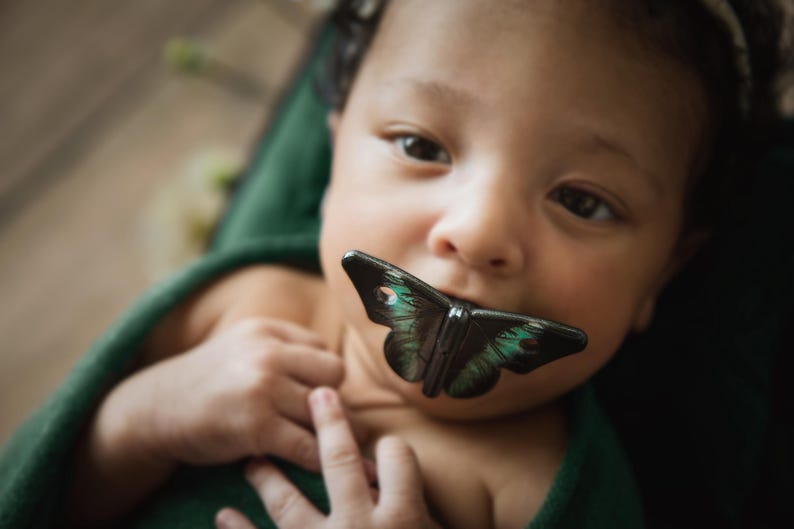 May include: A close-up of a baby's face with a black and green butterfly toy in their mouth. The baby is wearing a green shirt.