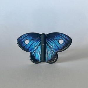 Puede incluir: Un amuleto en forma de mariposa azul y negro con una bisagra de metal negro. La mariposa tiene un dise&ntilde;o detallado con alas azules y negras.