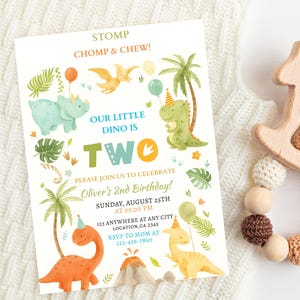 May include: A children's birthday invitation with dinosaur illustrations and the text "STOMP CHOMP & CHEW! OUR LITTLE DINO IS TWO." A wooden rocking horse-shaped rattle with a beaded ring is next to the invitation.