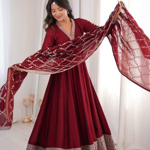 Puede incluir: Un vestido burdeos con una bufanda a juego. El vestido presenta un escote en V con bordados plateados y un dobladillo detallado. La bufanda tiene un patrón geométrico. El atuendo es adecuado para ocasiones especiales.