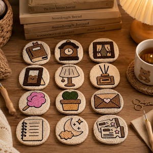 May include: A set of twelve circular, cream-coloured coasters featuring embroidered designs of a book, clock, window, picture frame, lamp, diffuser, candy floss, potted plant, envelope, notebook, moon, and tickets with brown and pink accents.
