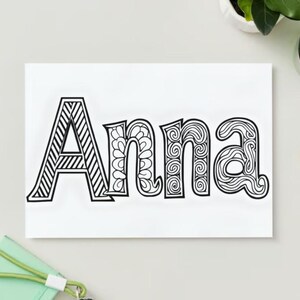 Op de afbeelding: Een wit rechthoekig canvas met de naam "Anna" in zwarte, decoratieve letters. De "A" heeft diagonale lijnen, terwijl de andere letters bloemen- en wervelpatronen hebben. Een potlood, een kleine plant en een groen notitieboek zijn ook zichtbaar.
