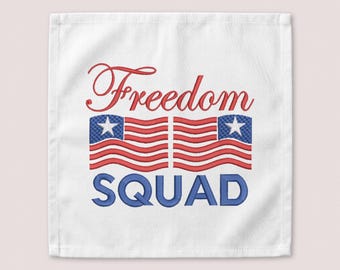 May include: A white square fabric towel featuring embroidered text that reads Freedom in a red cursive script above two stylised American flag motifs, with the word SQUAD embroidered in bold blue block letters at the bottom of the design.