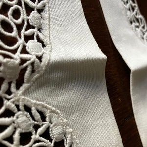 May include: Close-up of two white fabric strips with intricate floral lace edging. The fabric has a subtle texture, and the lace features delicate, openwork designs. The image highlights the contrast between the smooth fabric and the detailed lace.