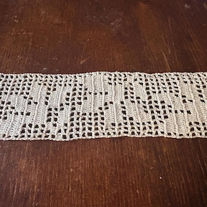 May include: A cream-colored, crocheted lace trim with a repeating geometric pattern. The trim is rectangular and appears to be made of a soft, natural fiber. The background is a dark brown wooden surface.