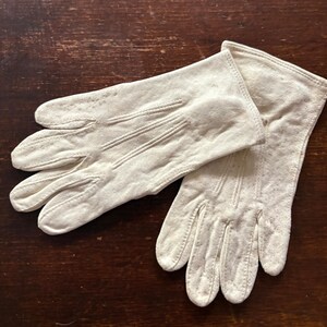 May include: A pair of off-white gloves featuring a soft, matte texture. Each glove has three vertical stitched lines running down the back of the hand, extending from the wrist toward the fingers, with a simple hemmed opening at the wrist.