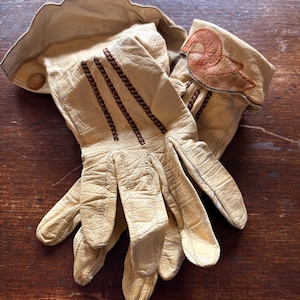 May include: A pair of vintage tan leather gloves. The gloves have brown stitching details and a decorative design on the cuff. The gloves are made of soft, supple leather and appear to be well-worn.