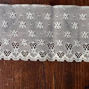 May include: Cream-colored lace trim with a floral pattern. The lace features a delicate openwork design with small, repeating flower shapes. The trim has a scalloped edge and is displayed against a dark wood surface.