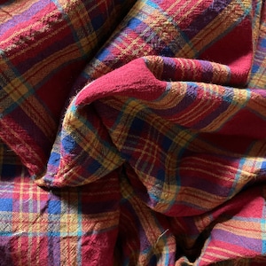 K&ouml;nnte beinhalten: Roter, blauer und gelber karierter Stoff mit einer strukturierten Oberfl&auml;che. Der Stoff ist gefaltet und drapiert, wodurch Muster und Textur sichtbar werden.