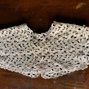 May include: Ivory-colored lace collar with a scalloped edge and intricate floral patterns. Small pink beads are interspersed throughout the design, adding subtle color. The collar is likely vintage or handmade.