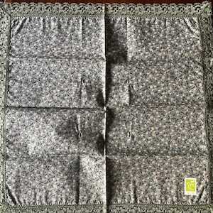 Puede incluir: Un cuadrado de tela floral verde y blanco con un borde de encaje verde. La tela tiene un pequeño patrón floral y una etiqueta verde con el texto "Lily & Co."