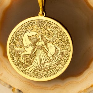 May include: A gold-colored pendant necklace featuring a detailed circular design. The pendant depicts a woman with a horse, moon, and stars. The necklace has a delicate chain and is displayed on a light-colored, patterned surface.