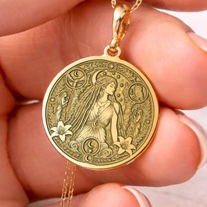 Könnte beinhalten: Eine goldfarbene Halskette mit einem detaillierten, kreisförmigen Anhänger. Der Anhänger zeigt eine Frau mit langen Haaren, umgeben von floralen und himmlischen Designs. Die Kette wird von einer Hand gehalten, die die feine Kette und den Anhänger zeigt.