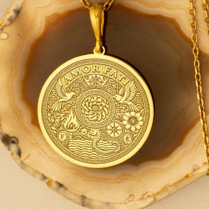 May include: A gold pendant necklace with a detailed circular design. The pendant features the words "AMOR FATI" and various symbols, including birds, flowers, and waves. The necklace is displayed on a light brown and white surface.