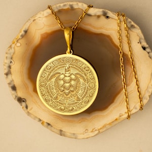 Puede incluir: Un collar con colgante dorado con un diseño detallado de tortuga. El colgante circular cuelga de una fina cadena dorada. La tortuga es el foco central, rodeada de intrincados patrones. Una joya llamativa.