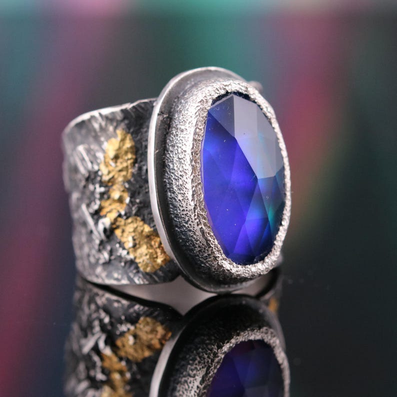 May include: A silver ring with a large, oval, faceted blue gemstone. The ring has a textured band with gold accents. The gemstone reflects light, showcasing its deep blue color and intricate facets. The ring is displayed on a reflective surface.