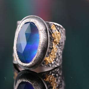 May include: A silver ring with a large, oval-cut blue gemstone. The band has a textured, dark finish with gold accents. The gemstone reflects shades of blue and purple, creating a striking visual effect. The ring is displayed on a reflective surface.