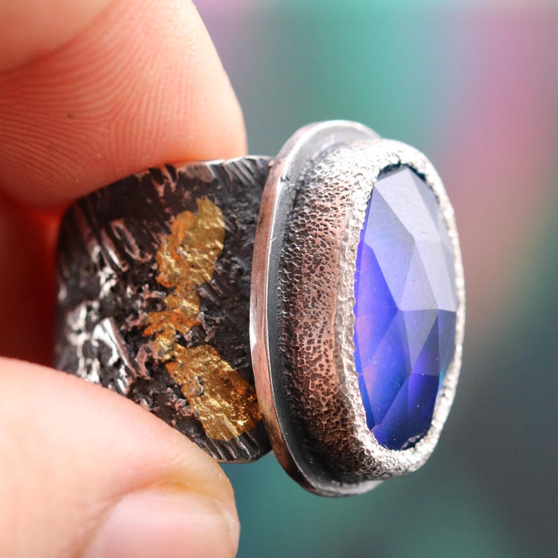 May include: A close-up of a silver ring with a large, faceted, oval-shaped blue gemstone. The ring has a textured band with gold leaf accents. The setting around the gemstone is also textured.