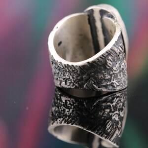 May include: Close-up of a silver ring with a textured band and a large, oval-shaped stone. The ring's band has a rough, bark-like texture, and the stone is set in a polished silver bezel. The ring is displayed on a reflective surface.