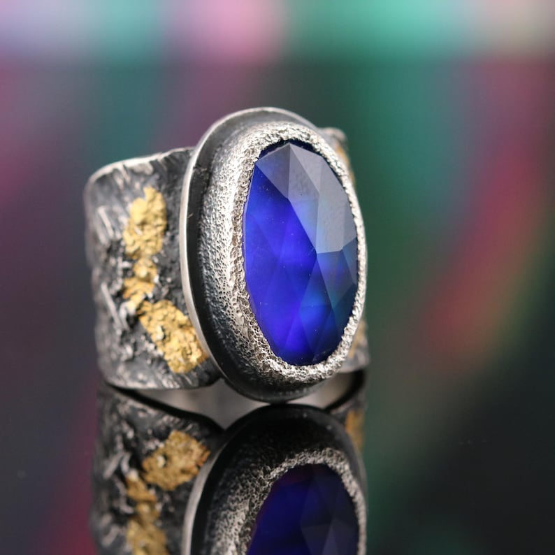 May include: A silver ring with a large, faceted, oval-shaped blue gemstone. The band has a textured, rustic appearance with gold-colored accents. The ring is displayed on a reflective surface.