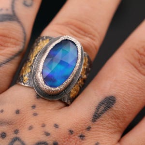 May include: A silver ring with a large, oval-shaped, faceted blue gemstone. The band has a textured design with gold accents. The ring is shown on a hand with tattoos.