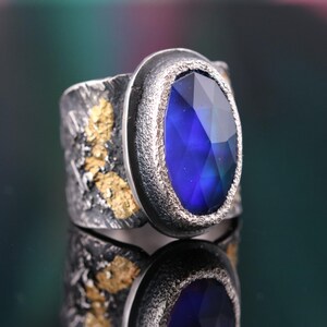 May include: A silver ring with a large, oval-cut, deep blue gemstone. The band has a textured, dark finish with gold-colored accents. The gemstone is set in a silver bezel, creating a striking contrast. The ring's design is bold and artistic.