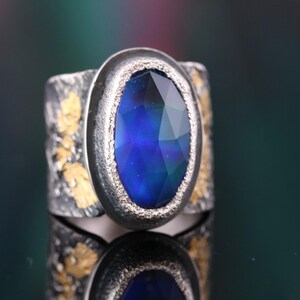 May include: A silver ring with a large, oval-shaped, faceted blue gemstone. The ring has a textured band with gold-colored accents. The gemstone is set in a silver bezel, creating a striking contrast. The ring is displayed on a reflective surface.