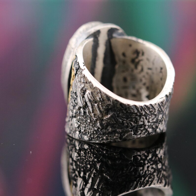 May include: Close-up of a silver ring with a textured band and a smooth, oval-shaped stone. The ring has a rustic, handcrafted appearance with a gold accent near the stone. The ring is displayed on a reflective surface.