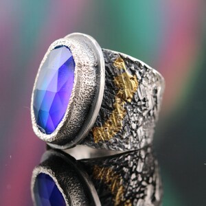 May include: A silver ring with a large, oval-shaped, iridescent blue gemstone. The band has a textured, dark gray finish with gold accents. The ring is displayed on a reflective surface, showcasing its design and color.