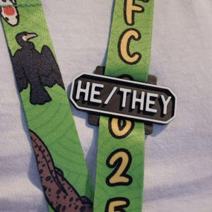 May include: A green lanyard with a black and white badge that says "HE/THEY". The lanyard has a pattern of birds, fish, and a spotted animal.