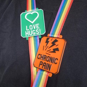 May include: Two rectangular badges with rainbow ribbons. One is green with a white heart and the words "LOVE HUGS!". The other is orange with black lightning bolts and the words "CHRONIC PAIN".
