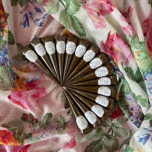 Peut inclure: Un ensemble de cônes de henné bruns, disposés en éventail. Chaque cône est orné d'une étiquette blanche avec un motif stylisé. L'ensemble est présenté sur un tissu à motifs floraux roses, blancs et verts.