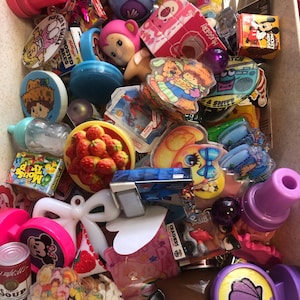May include: A collection of vintage toys and trinkets in a box. Items include a pink bottle stopper, a blue bear, a pink bear, a Mickey Mouse box, and a can of soup. Other items include a strawberry-shaped item, and a yellow and blue toy.