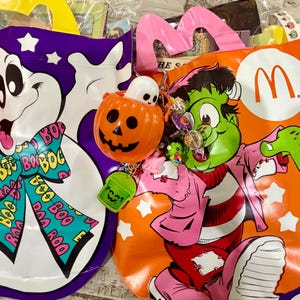 May include: Two plastic Halloween bags featuring a white ghost on a purple background and a green Frankenstein monster on an orange background with the McDonald's logo. Each bag is decorated with a colorful bow and small orange pumpkin buckets.